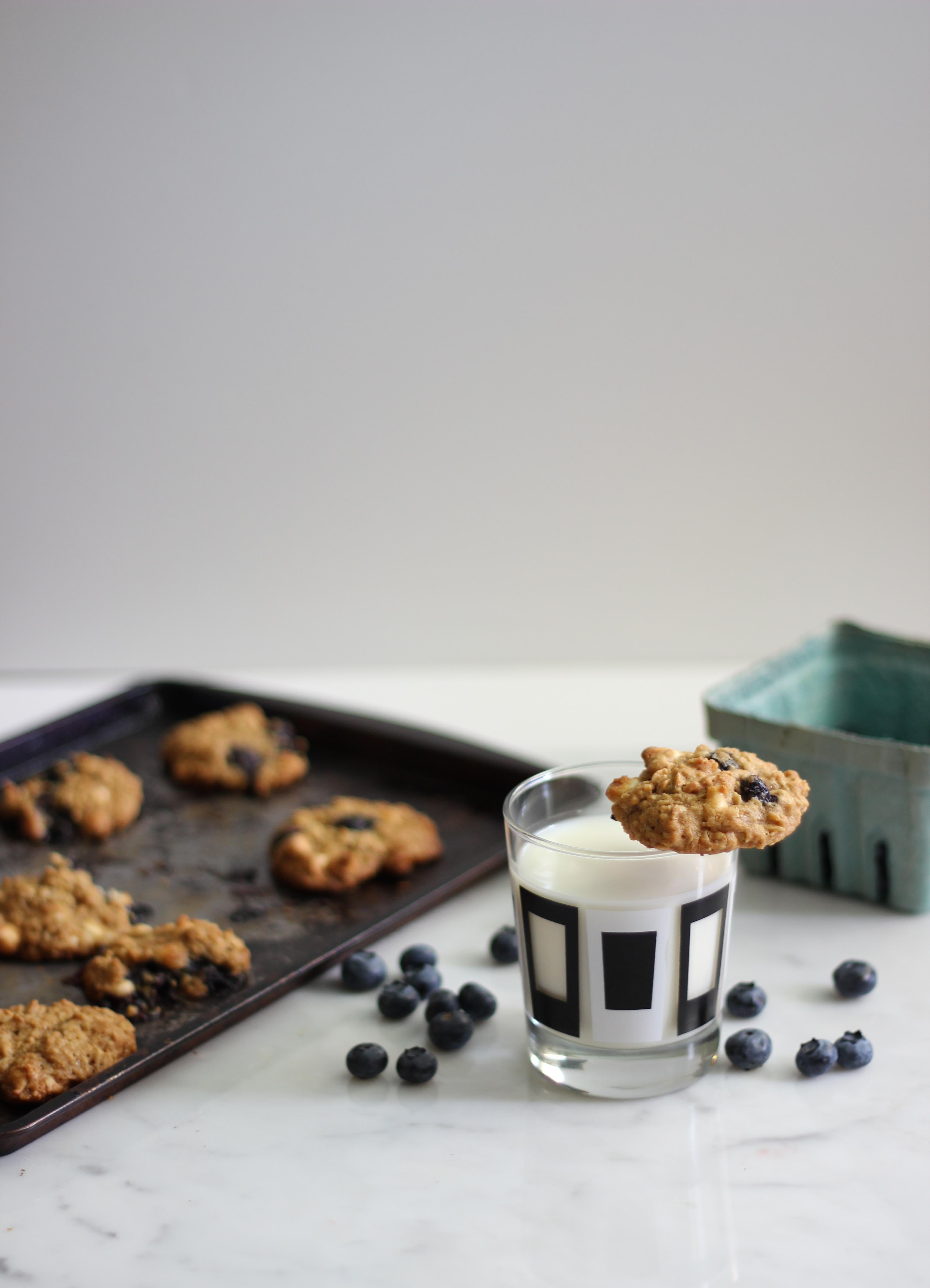 blueberry oatmeal cookies on a baking sheet with one cookie perched on top of a glass of milk.
