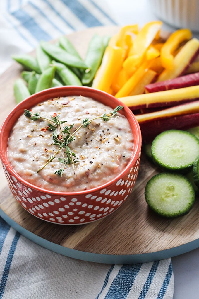 Lemon Herb White Bean Dip in a small orange bowl on a board next to cut vegetables. 