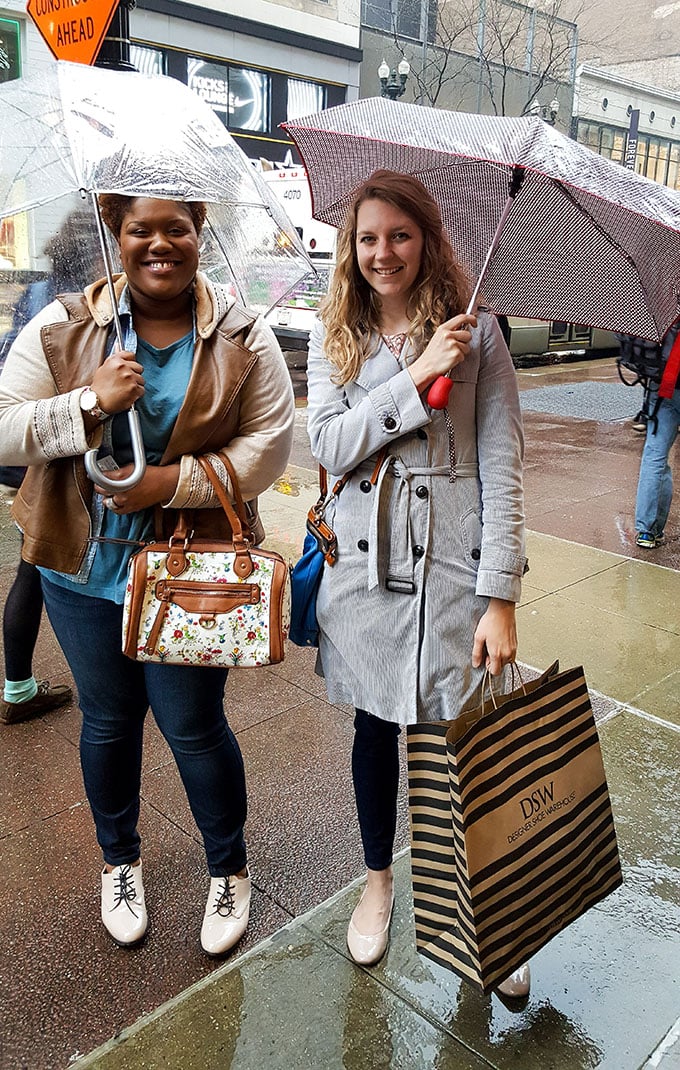 two women standing on the sidewalk smiling and holding umbrellas. 