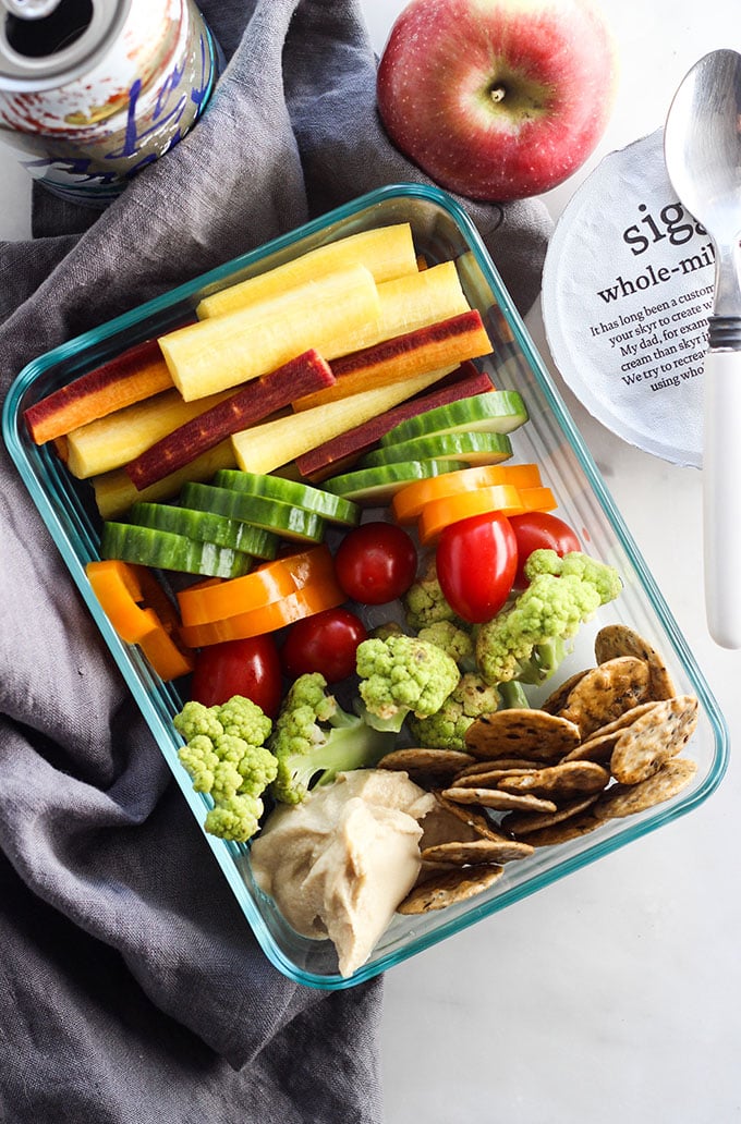 Glass meal prep container with colorful vegetables, hummus, and crackers. 