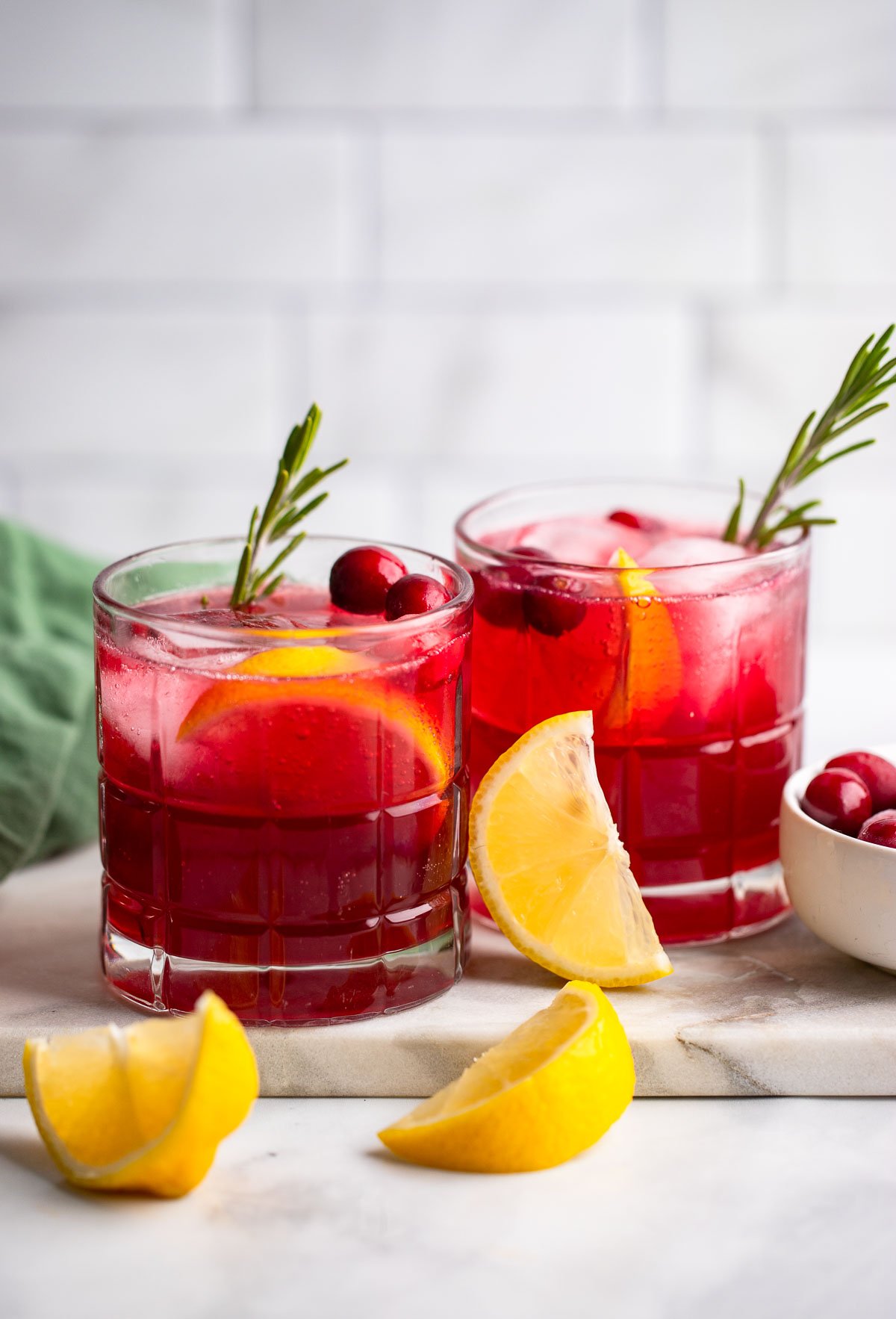 two glasses of cranberry cocktail garnished with lemon, rosemary, and fresh cranberries.