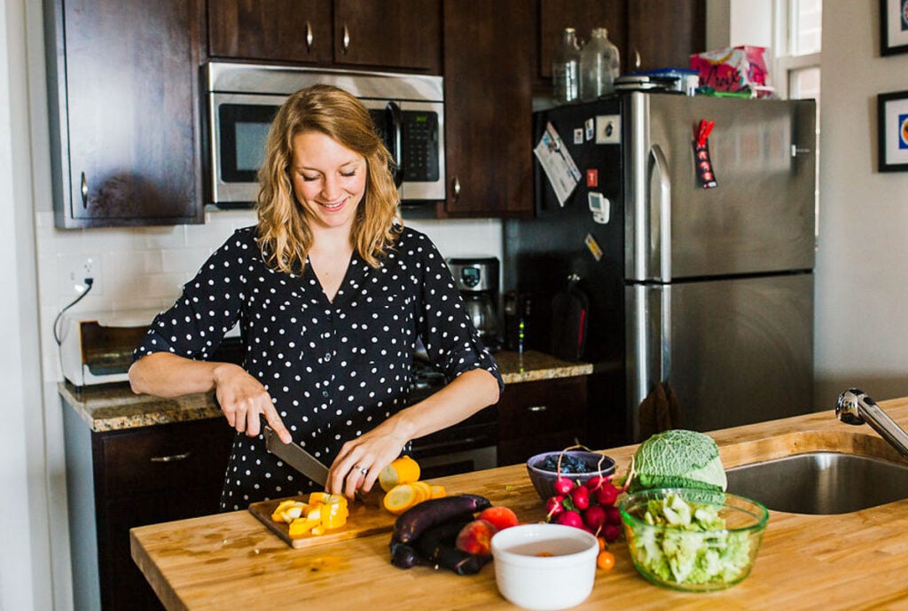 blonde woman standing in kitchen chopping vegetables. 