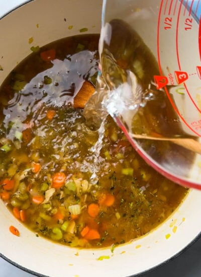 broth pouring from measuring cup into pot with chicken noodle soup.