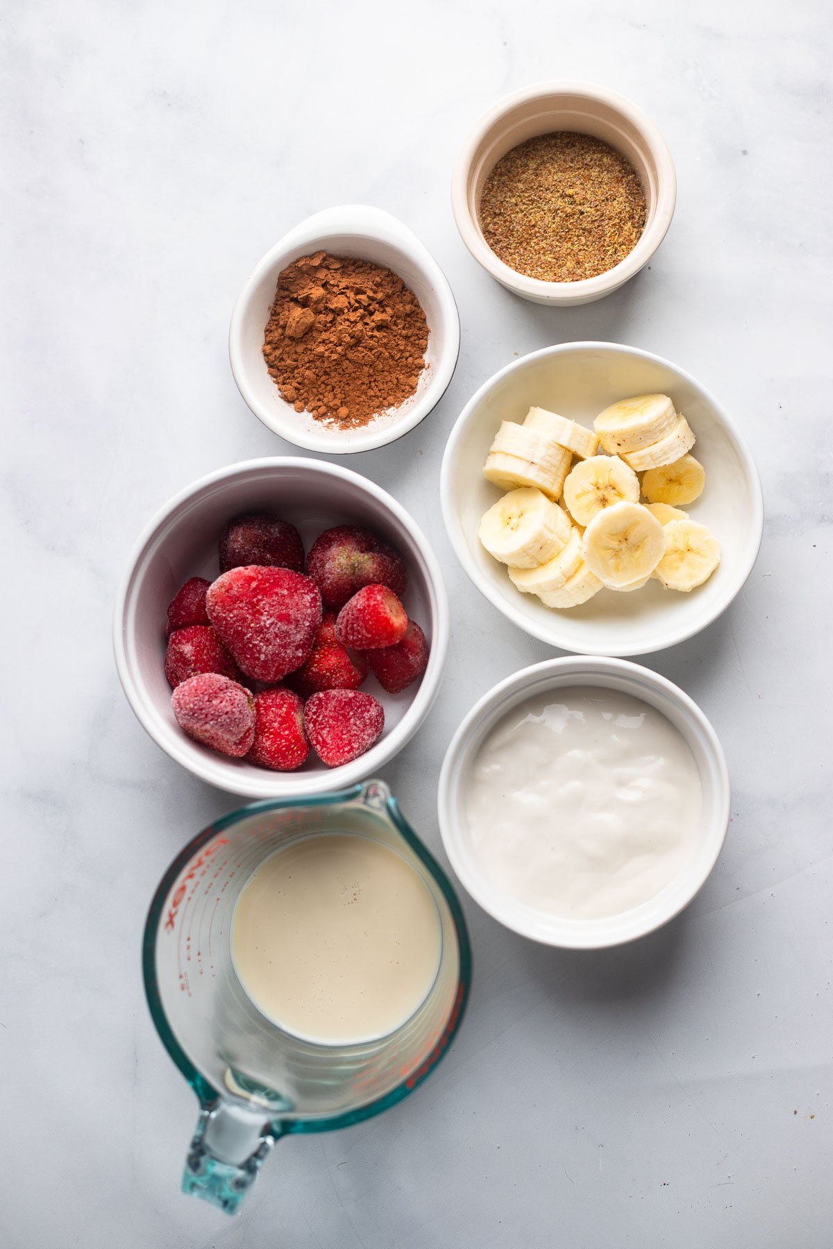 chocolate strawberry banana smoothie ingredients in small bowls laid out on marble backdrop. 