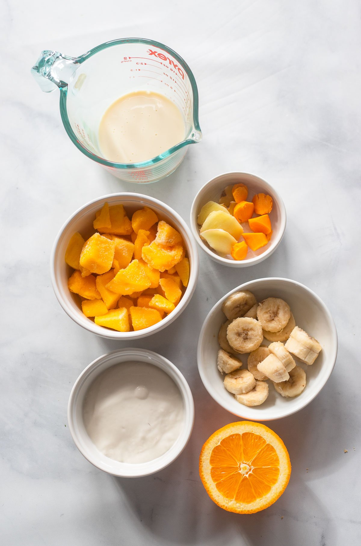 mango turmeric smoothie ingredients in small bowls set out on a marble backdrop.