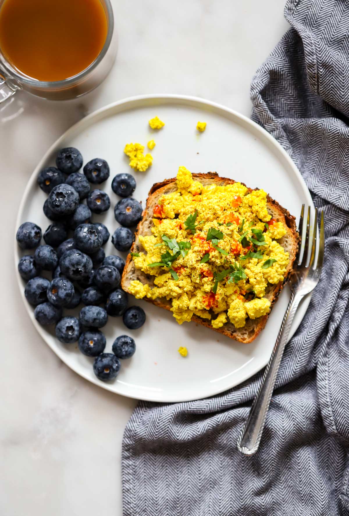 vegan tofu scramble piled on a piece of buttered toast sitting on a plate with some fresh blueberries. 
