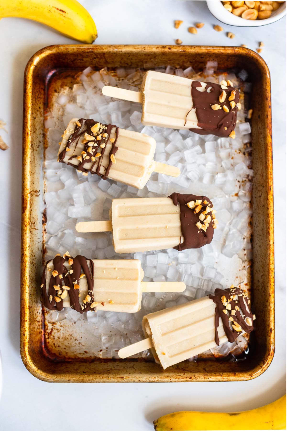 peanut butter banana popsicles dipped in chocolate on a small sheet pan with ice.