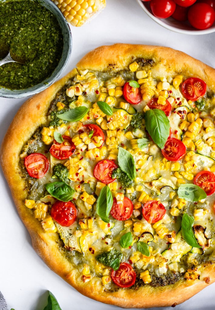 close up of summer vegetable pizza topped with corn, pesto, tomatoes, and fresh basil. 