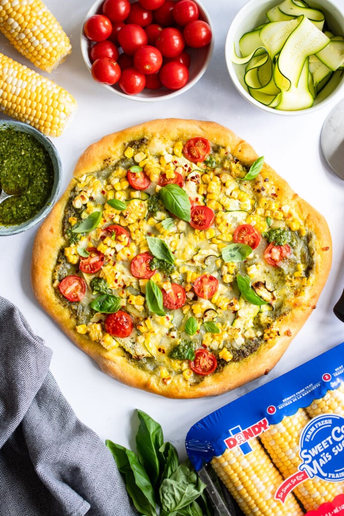 sweet corn pizza garnished with basil on white background surrounded with bowls of pizza ingredients.